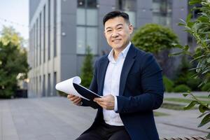 Successful young asian man businessman sitting on a bench during a work break near the office center and holding a folder with papers and documents in his hands, looking and smiling at the camera. photo