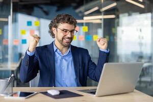 Mature experienced businessman in business suit is looking at successful achievement results, boss with laptop inside office holding hands up, gesture of success, triumph, online notification laptop. photo