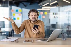 A puzzled businessman at his office desk, looking frustrated while using his smartphone, surrounded by sticky notes and a laptop. photo