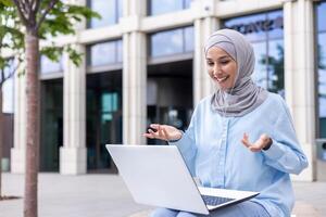 sonriente musulmán mujer en un ligero azul hijab comprometido en un animado llamada en su computadora portátil, sentado fuera de un moderno edificio. foto