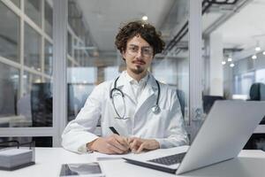 un médico en un blanco Saco y estetoscopio es visto sentado a un escritorio, enfoque en su ordenador portátil. hombre grave y pensando escritura reporte foto