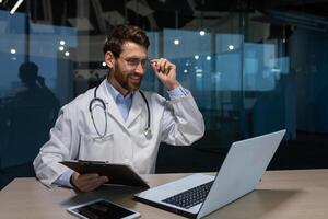 A young male doctor is talking to a patient using a call from a laptop. He is sitting at a table in an office in a hospital. He holds a folder with documents, adjusts his glasses, talks, smiles. photo