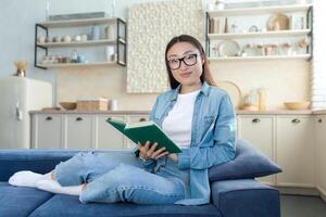 Young beautiful Asian woman in denim clothes and glasses sitting on sofa at home. He holds a green book in his hands, looks at the camera, smiles. Rest for the weekend. photo