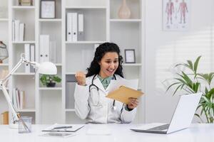 A cheerful mature female doctor excitedly celebrates while reading good news from an envelope in her bright, organized clinic office, exuding professionalism and happiness. photo