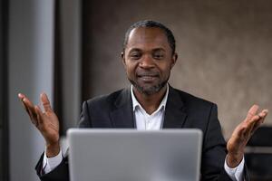 A mature African businessman in a suit talks and gestures in front of a laptop during an online meeting. He appears engaged and proactive. photo