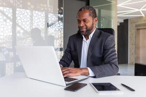 exitoso maduro africano americano empresario a trabajo dentro oficina, experimentado jefe trabajando con computadora portátil, hombre sonriente satisfecho con logro resultados. foto