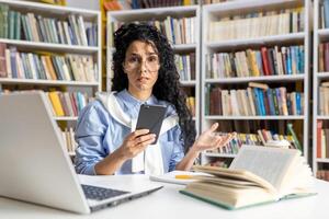 un perplejo Hispano mujer multitarea con un teléfono inteligente y ordenador portátil en un biblioteca, rodeado por libros, expresando preocupación y confusión. foto