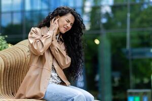 Frustrated woman sitting on modern curved bench outdoors. Disappointment female holding hand to ear and feeling discomfort or pain in that area. Background of building with large glass windows. photo