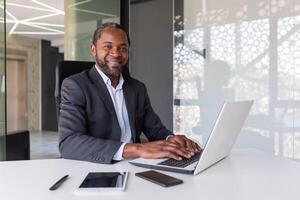 retrato de exitoso contento africano americano jefe, hombre sonriente y mirando a cámara, empresario en negocio traje sentado a escritorio con ordenador portátil dentro oficina. foto