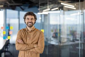 Confident mature Hispanic male with a warm smile standing casually in a modern office environment, exuding friendly professionalism. photo