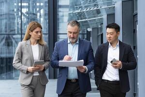 Diverse business team three colleagues employees discuss documents contract outside office building businessman and businesswoman holding bills and financial reports talking photo