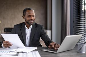 Confident African American businessman in formal attire working on a laptop while reviewing documents at his office desk. photo