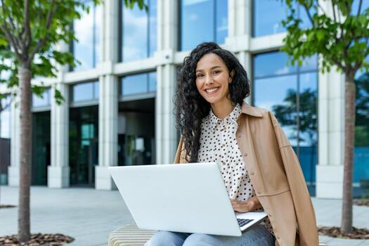 alegre Hispano mujer sentado al aire libre con un computadora portátil, sonriente como ella trabajos en un moderno urbano ajuste. ella es vistiendo un elegante Saco y se sienta por casualidad. foto
