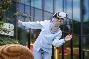 un sonriente mujer en un hijab disfruta un virtual realidad auriculares experiencia fuera de un moderno edificio con un alegre expresión. foto