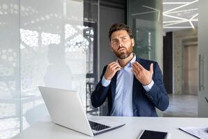 Confident businessman in office adjusting tie, laptop on desk. Concept of leadership, professionalism, and corporate lifestyle. photo