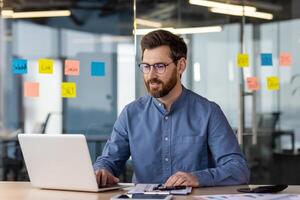 Successful and self-confident young businessman working concentratedly in the office behind a laptop. Sitting at the desk smiling, checking data and accounts, talking on a call. photo