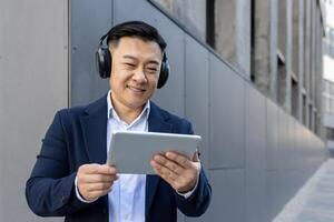 profesional masculino en un traje multitarea con un digital tableta y auriculares fuera de un moderno edificio. foto