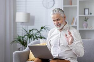 Elderly man relaxing at home, using a laptop and greeting someone with a wave in a modern living room setting. photo