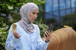 Upset cheated woman with phone trying to make online purchase and money transfer, muslim woman in hijab disappointed got rejection, holding phone and bank credit card, dissatisfied buyer. photo