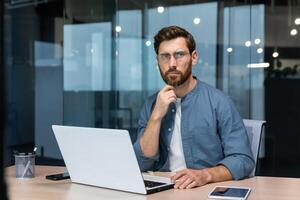 .Serious pensive businessman in shirt thinking about decision sitting at table in modern office, man with beard is using laptop at work. photo