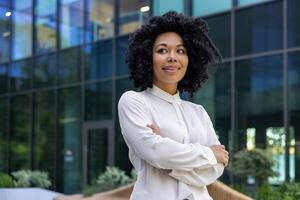 Confident adult mature businesswoman outside urban office business building, African American boss with crossed arms looking away, female worker in shirt satisfied with achievement results at work. photo