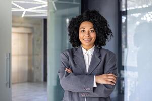 Portrait of successful female boss in business suit, mature African American woman smiling and looking at camera with crossed arms, businesswoman happy with achievement at workplace inside office. photo