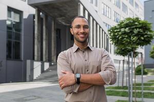 retrato de joven Hispano estudiante hombre sonriente y mirando a cámara cerca Universidad instalaciones con brazos cruzado vistiendo lentes y camisa foto