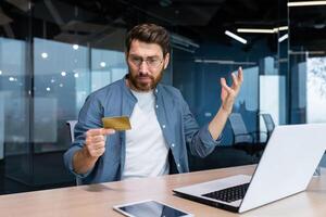 engañado y decepcionado empresario trabajando dentro oficina, hombre participación banco crédito tarjeta y molesto a hacer un compra en en línea almacenar, en línea fraude y engaño. foto