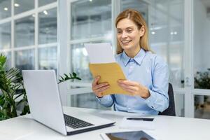 sonriente profesional mujer leyendo un carta, expresando alegría y satisfacción a su oficina espacio de trabajo foto