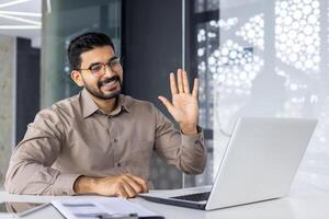 un alegre masculino profesional en un inteligente camisa olas calurosamente a su ordenador portátil durante un virtual reunión en un moderno oficina ajuste. foto