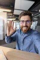 Vertical close-up photo of a young smiling man standing in a business office wearing glasses and talking on a call, greeting and waving at the camera.