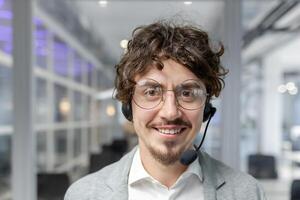 Smiling young businessman with curly hair wearing a headset in a modern office. Energetic male professional busy at work. photo