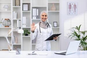 Smiling Muslim doctor in hijab greeting with a wave while holding a clipboard in a well-lit medical office. photo