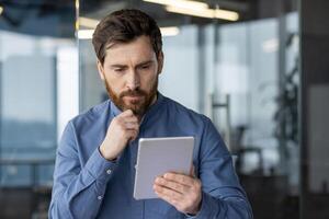 Close-up photo of a young man in a blue shirt who is seriously sitting at the desk in the office and thoughtfully looking at the screen ,of the tablet he is holding in his hands.