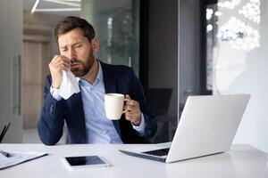 A young man with a cold does not feel well, works in the office, sits at the table in a suit, holds a cup of hot drink with medicine in his hand, wipes his nose with a napkin. photo