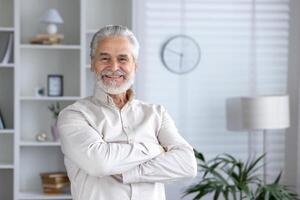 retrato de un alegre mayor hombre en pie con brazos cruzado a hogar, radiante confianza y positividad en un cómodo, elegante ajuste. foto