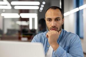 A close-up portrait of a young African American man sitting in the office in front of a laptop screen, intently and focused on work, a project, thinking about a solution to a problem. photo