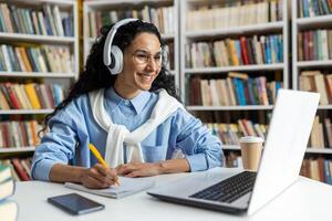 enfocado estudiante se involucra con en línea aprendizaje en un biblioteca configuración, utilizando auriculares y tomando notas junto a un taza de café. foto