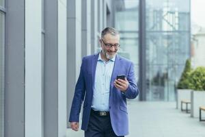 Senior gray-haired handsome man in a suit and glasses. Walks down the street, holds the phone in his hands, makes a call, talks. photo