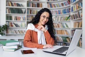 joven Hispano mujer estudiando en académico Universidad biblioteca, hembra estudiante sonriente y mirando a cámara mientras sentado a computadora portátil, mujer con Rizado cabello. foto