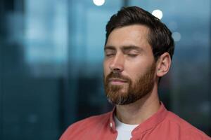 Close up of mature thinking businessman meditating and thinking with closed eyes at workplace inside office, man in red shirt resting and breathing. photo