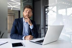 Pensive businessman contemplating at desk with laptop in a modern office setting, portraying deep thought and professionalism. photo