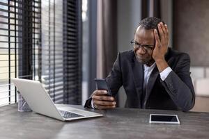 Stressed african american entrepreneur grabbing head with hand while using smartphone in remote workspace. Frightened man getting warning notification from banking application while sitting by desk. photo