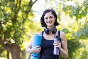 A smiling woman equipped with yoga mat, headphones, and water bottle enjoying the natural outdoor setting for her fitness routine. photo