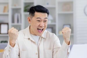 de edad mediana asiático hombre en un crema camisa alegremente celebrando mientras mirando a su ordenador portátil pantalla, evidentemente emocionado por bueno Noticias o éxito a hogar. foto