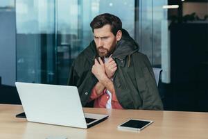 es frío en el oficina, el empresario es congelación mientras trabajando en un ordenador portátil dentro el edificio, el hombre es cubierto con un calentar chaqueta. foto