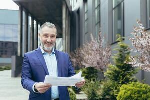 Real estate agent. A handsome senior man and beard stands on the street near the houses, office center, holding documents in his hands, satisfied with a successful deal. Looks at the camera, smiles. photo