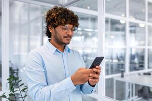 un joven indio hombre es en pie en el oficina y utilizando el teléfono, discado mensajes, enviar mensajes de texto, en línea pago, ordenando, compras. foto