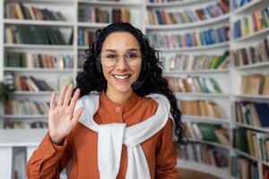 Joyful curly Hispanic girl sitting at a table in front of a laptop in a headset looking at the camera and waving at the camera, office worker in a call center, working call camera view. photo