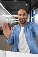 A young, cheerful businessman greets colleagues with a wave during a virtual meeting in a modern office setting. photo